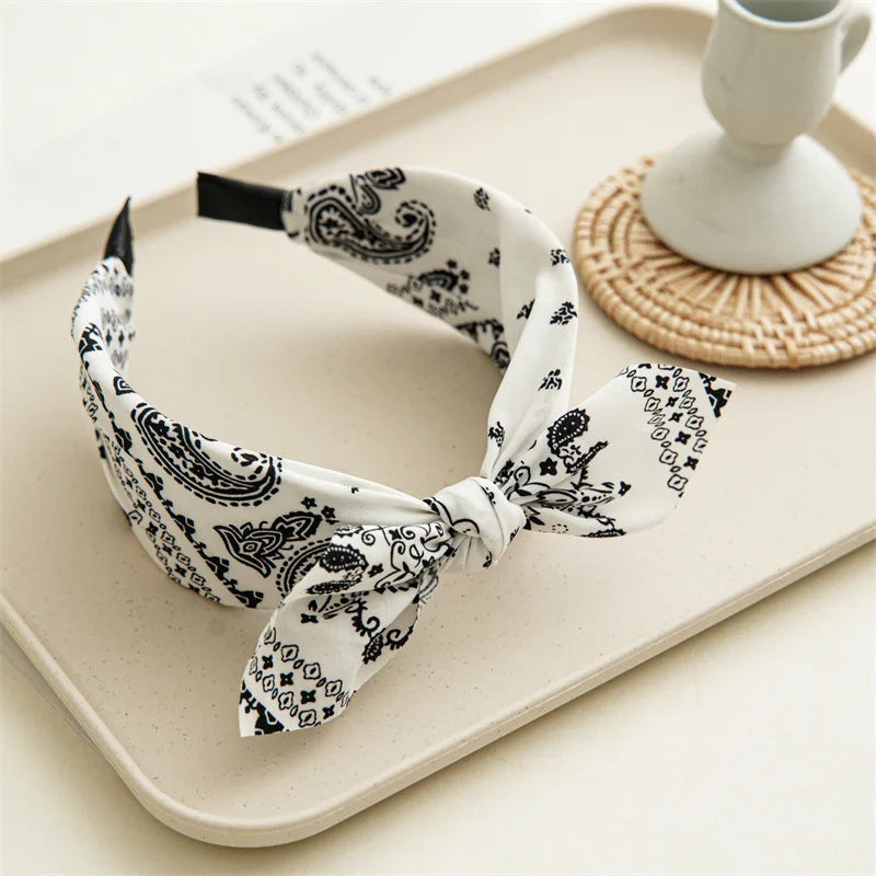 A black and white paisley bandana headband tied in a bow, displayed on a light tray next to a white cup.