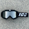 A pair of black motorcycle goggles with a reflective lens and a white '100' marking on the strap, resting on a gray surface.