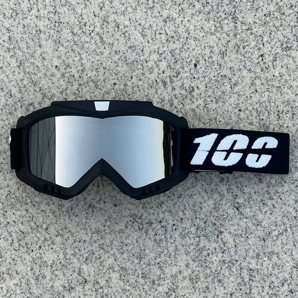 A pair of black motorcycle goggles with a reflective lens and a white '100' marking on the strap, resting on a gray surface.