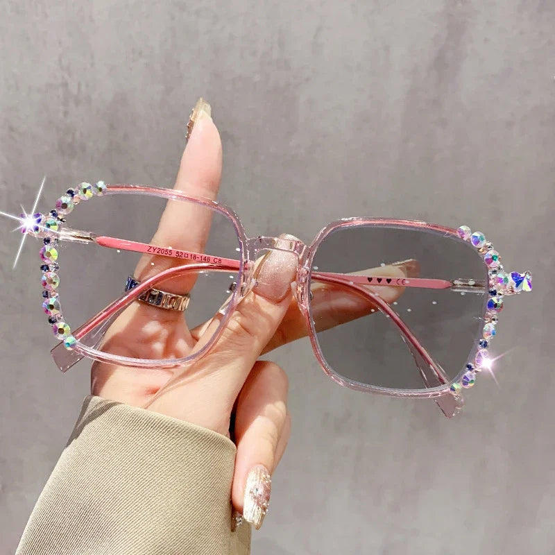 A hand holds a pair of pink photochromic diamond style eyeglasses with sparkling accents against a gray background.