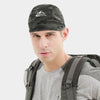 Un hombre vistiendo una gorra de camuflaje con logo de montaña, camiseta gris y mochila.