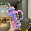 A hand holds a purple glass reusable water bottle with a straw and handle, filled with liquid.