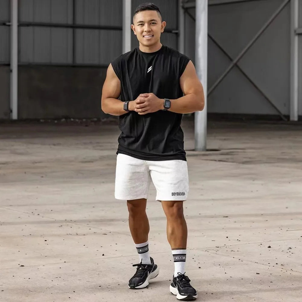 A man wearing a black sleeveless fitness tank top and white shorts posing indoors.