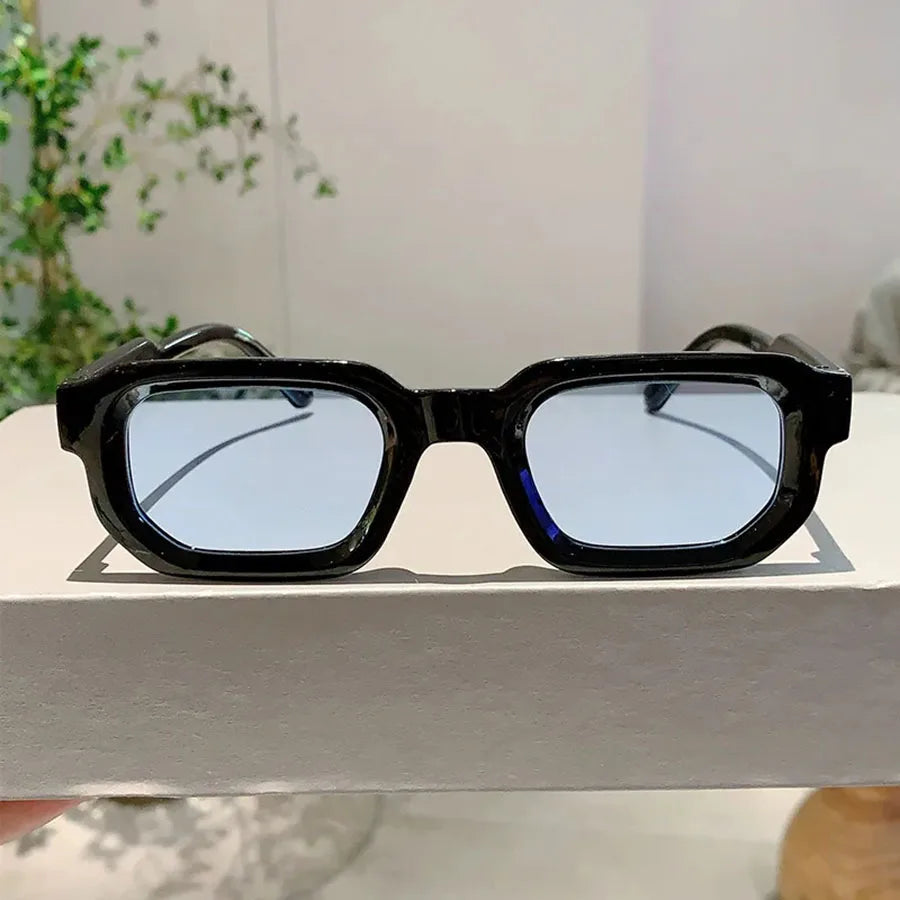 Lunettes de soleil à monture rectangulaire noire rétro avec des verres miroirs bleus posées sur une surface blanche.