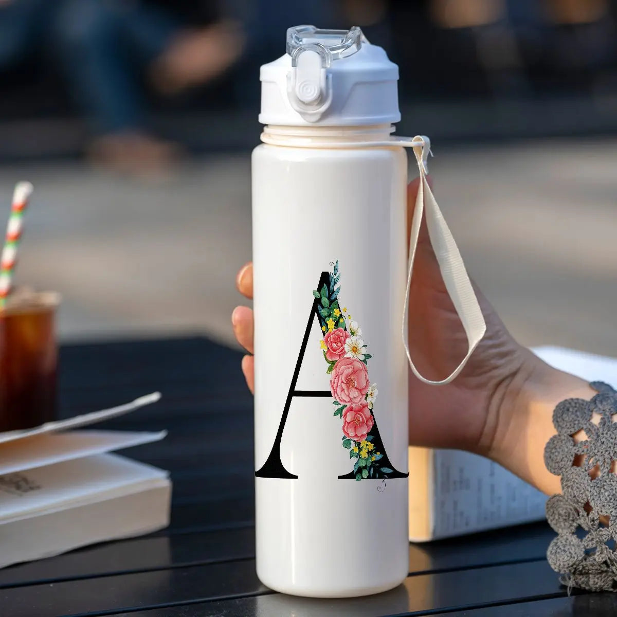 A white insulated tumbler with a black letter A design is held in a hand on a dark surface.
