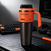 An orange insulated stainless steel tumbler with a handle sits on a dark desk next to a laptop and keyboard.