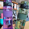 A hand holds a clear purple water bottle with a black cap and a glass bottle with a green liquid and a black cap.