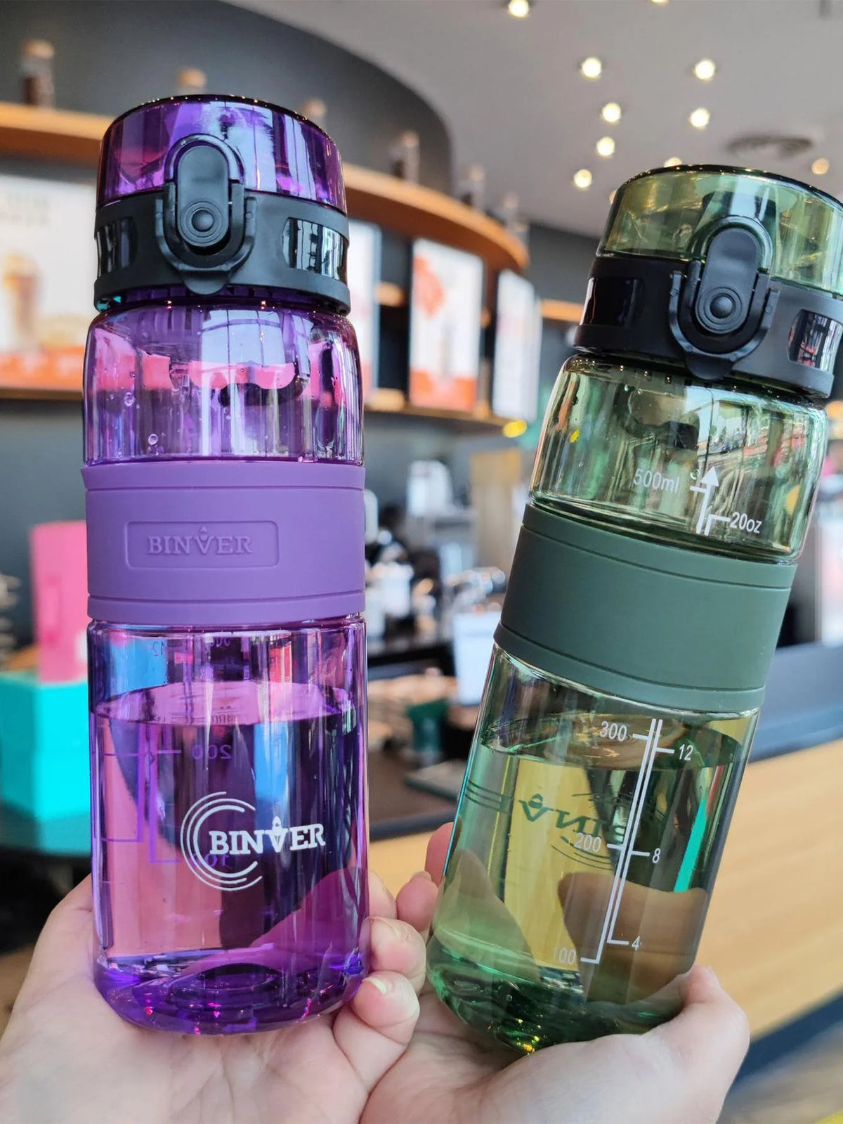 A hand holds a clear purple water bottle with a black cap and a glass bottle with a green liquid and a black cap.