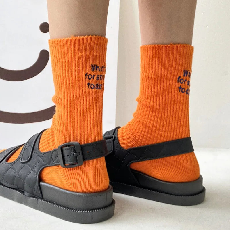 A pair of bright orange knitted socks with embroidered text are shown on a person's legs.