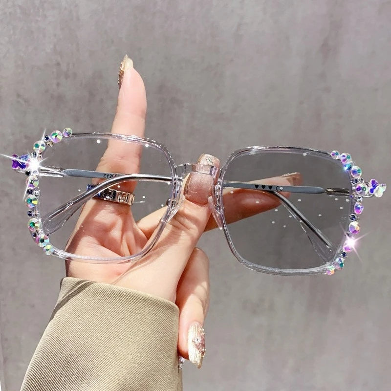 A hand holds a pair of clear photochromic diamond style eyeglasses decorated with sparkling gems.