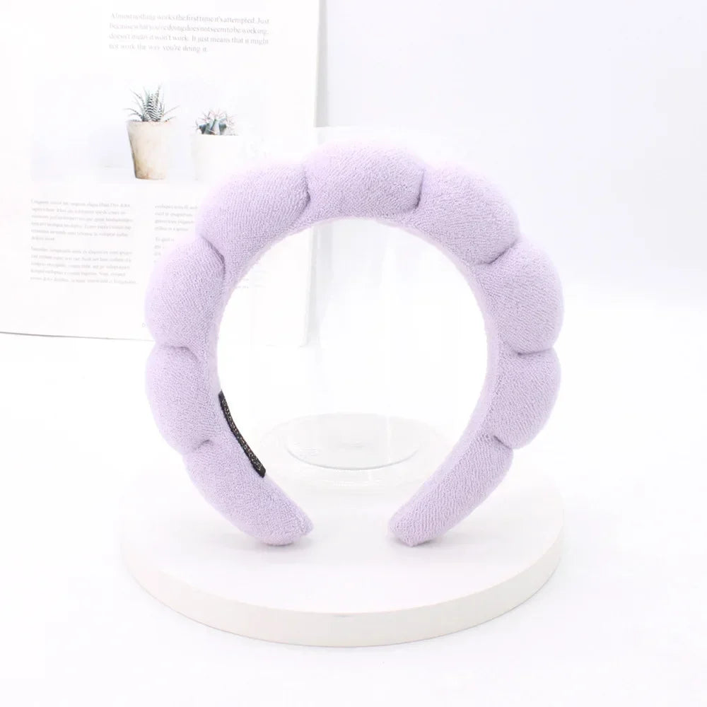 A lavender sponge terry cloth headband for hair styling displayed on a white circular stand.