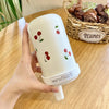Un tumbler isotherme blanc décoré de cerises rouges tenu par une main, posé sur une surface en bois.
