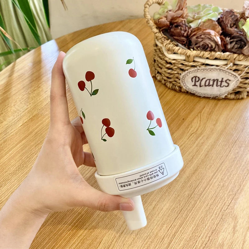 Un tumbler isotherme blanc décoré de cerises rouges tenu par une main, posé sur une surface en bois.