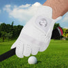 A hand wearing a white microfiber golf glove with a logo grips a white golf ball on green grass.