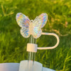 A clear silicone butterfly straw cover is shown resting on a glass straw, set against a blurred green outdoor background.