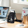A black neoprene water bottle carrier bag with a shoulder strap resting on a wooden table next to a laptop and papers.