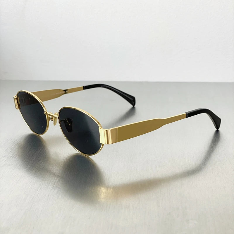 Gold-toned metal oval sunglasses with black frames and dark lenses resting on a gray surface.