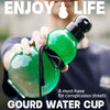Eine Hand hält eine grüne Kürbis-Wasserflasche mit schwarzem Trageriemen vor einem verschwommenen Hintergrund.