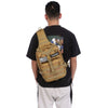 A man wearing a black t-shirt and light pants is shown from the back carrying a tan tactical sling backpack.