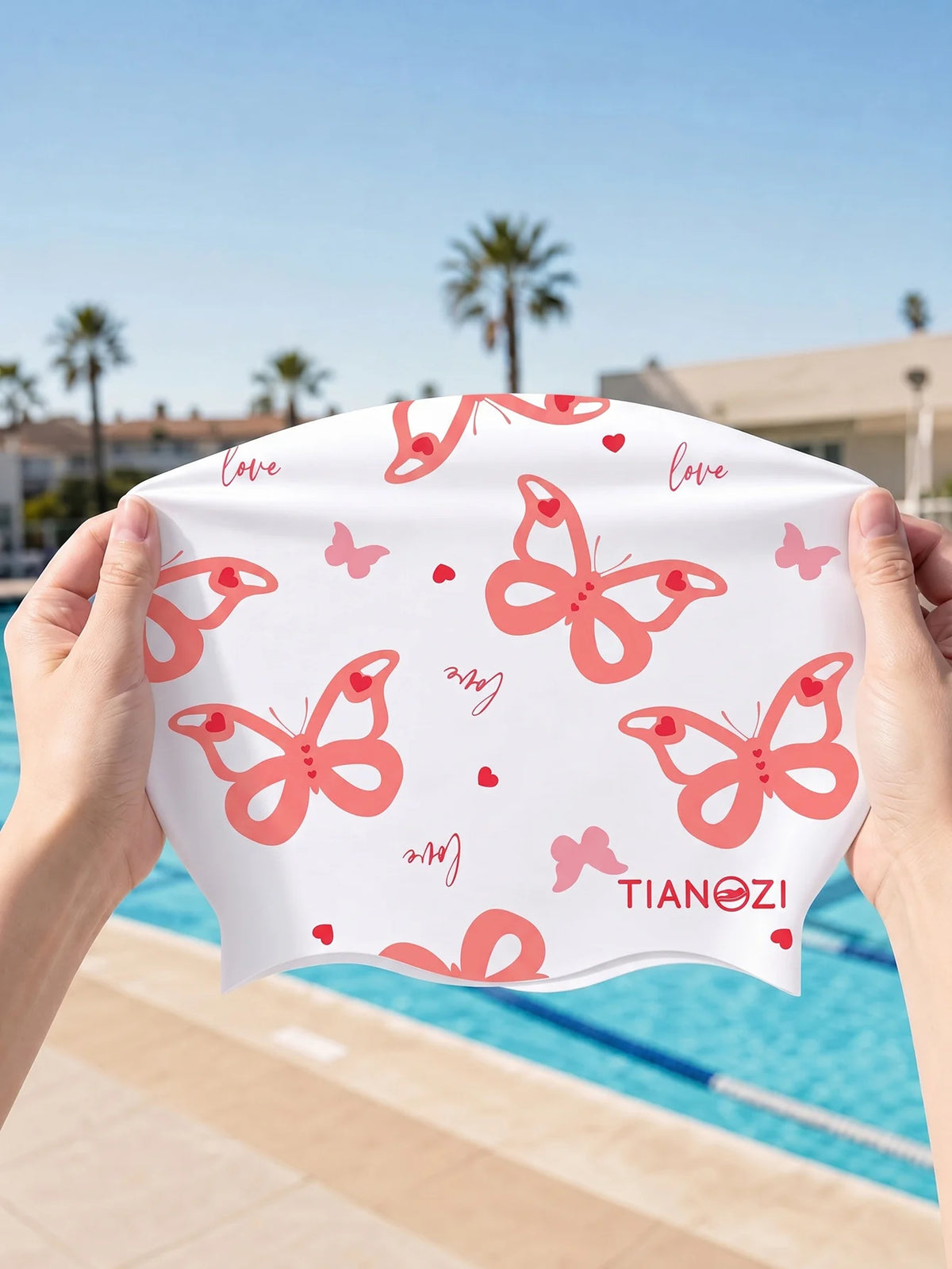 A hand holds a pink butterfly print silicone swimming cap against a bright blue pool background.