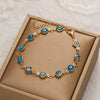 Gold charm bracelet with blue and white evil eye charms displayed in a light brown box.