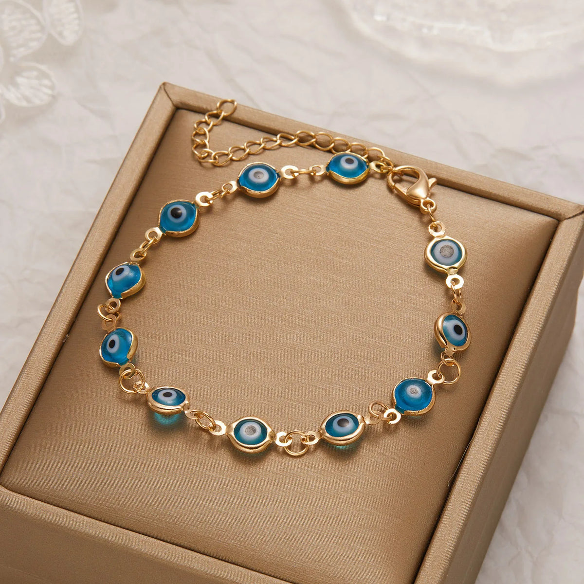 Gold charm bracelet with blue and white evil eye charms displayed in a light brown box.