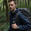 Mann mit dunkelblauer Jacke und Rucksack in Wald, hält eine Smartwatch.