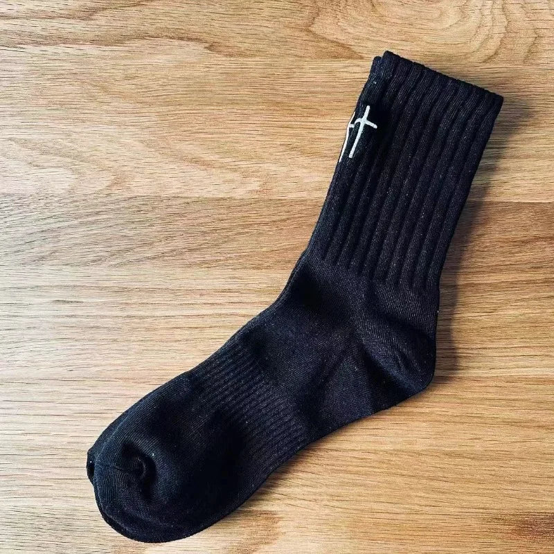 A black mid-length sock with a ribbed texture and a white cross design is laid on a wooden surface.