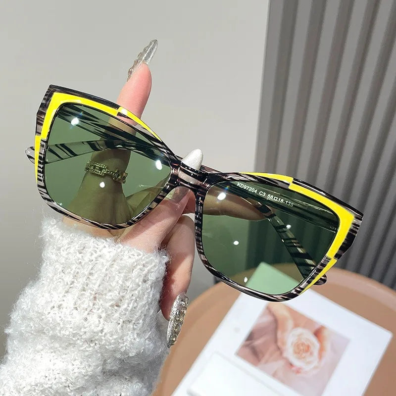 A hand holds a pair of vintage square cat eye sunglasses with yellow frames and green lenses against a light background.