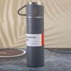 A tall, matte gray stainless steel vacuum flask with a handle and a red label indicating 12 hours cold.