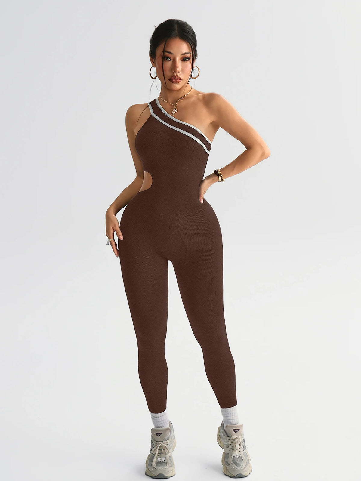 A model wearing a seamless, dark brown yoga jumpsuit with white trim and gold jewelry.