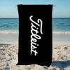 A black microfiber beach towel with white script text reading 'Tiltless' hanging on a wooden rod on a sandy beach.