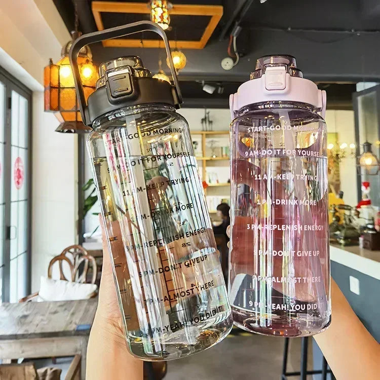 A hand holds a clear insulated water bottle with a pink lid and straw, showing various motivational text on the glass.