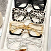 A display of four oversized rectangular reading glasses in black, leopard print, amber, and clear frames.