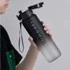 A hand holds a black sports water bottle with a black cap and a green tag against a light gray background.