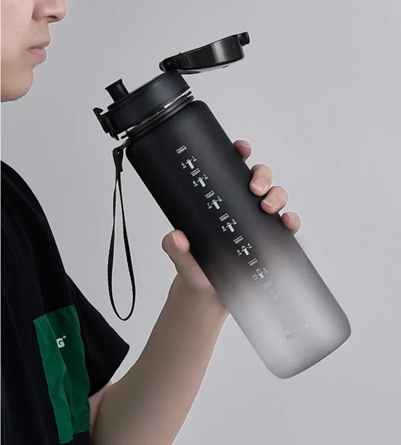 A hand holds a black sports water bottle with a black cap and a green tag against a light gray background.