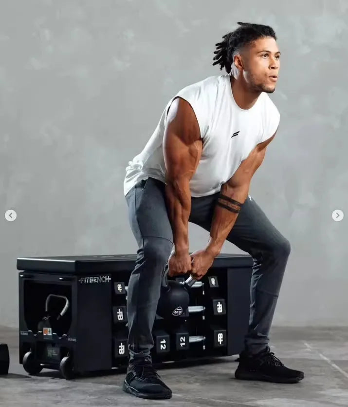 A muscular man wearing a white sleeveless fitness tank and dark pants is performing a lift with a black weight plate on a gym