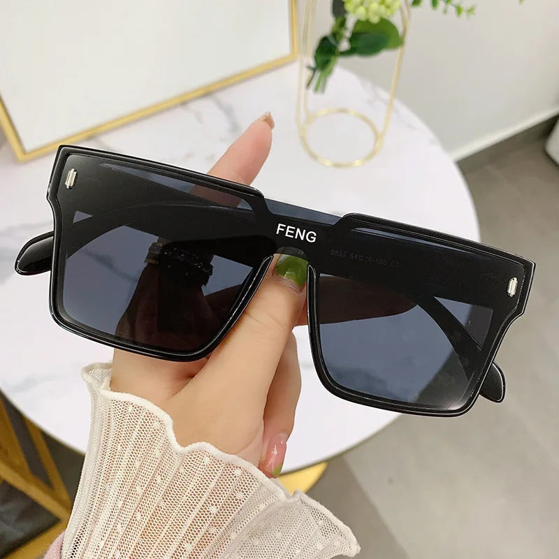 A hand holds a pair of black oversized square sunglasses with gold accents against a white background.