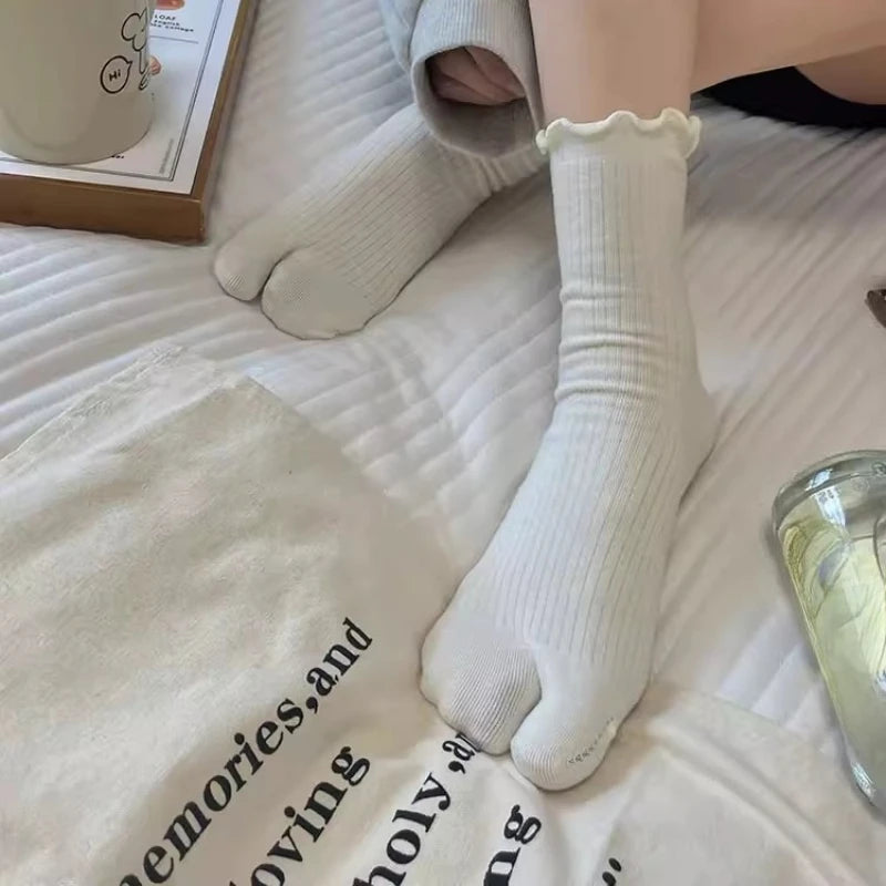 A close-up of white knit crew socks with ruffle cuffs worn on a foot, resting on a white fabric.
