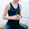 A woman holds a black protective water bottle sleeve with a phone holder against a blurred outdoor background.
