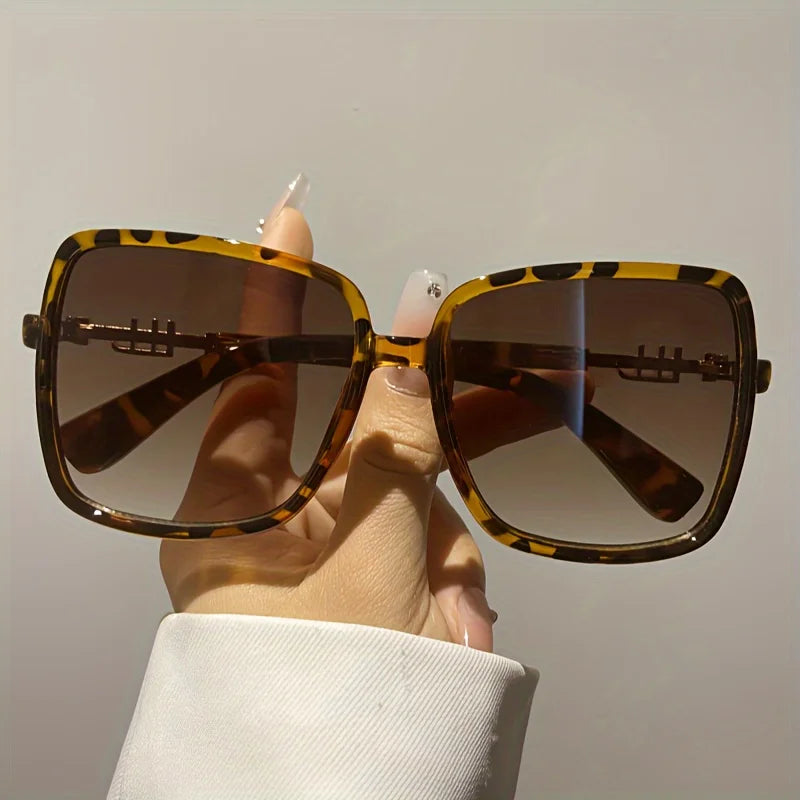 A hand holds a pair of retro two-tone gradient sunglasses with brown and black leopard print frames.