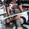 A person wearing black athletic gear is using neoprene knee pads while lifting weights in a gym setting.