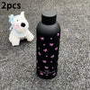 Dos pequeños juguetes de perros blancos y una taza térmica negra con diseños de mariposa rosa sobre una superficie gris.