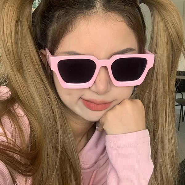 A young woman models pink retro square frame sunglasses with dark lenses against her long blonde hair.