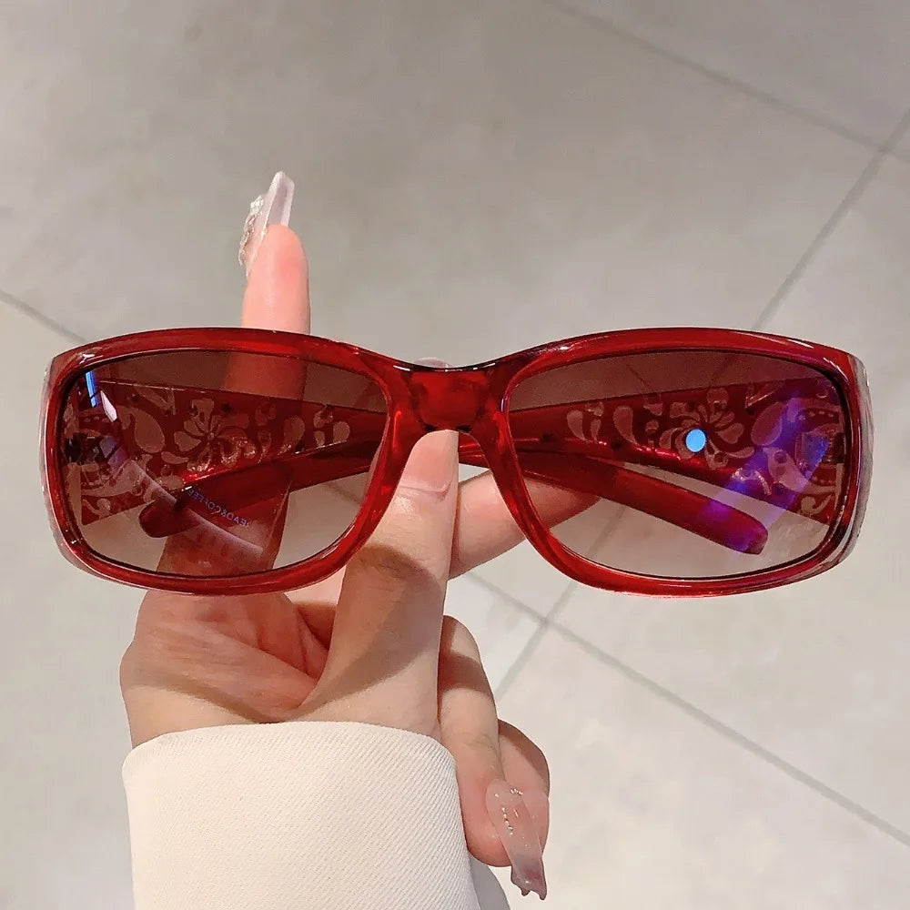 A hand holds a pair of red tinted Y2K cat eye rhinestone sunglasses against a light background.