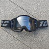 Une paire de lunettes de moto tout-terrain et ATV noires posées sur une surface grise texturée.