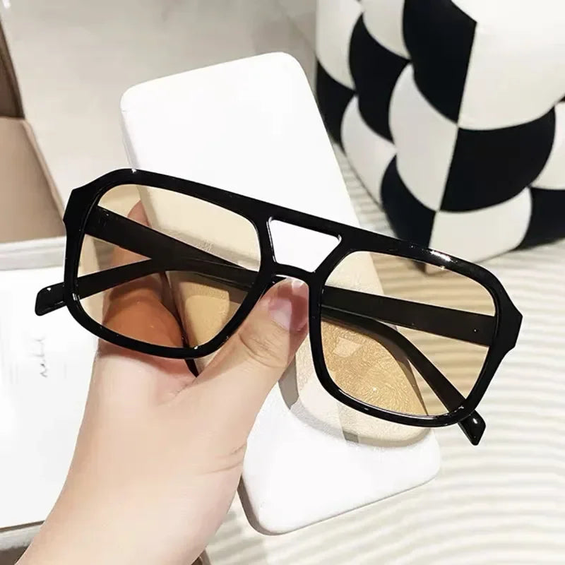 Un par de gafas de sol cuadradas con montura negra y lentes dorados sobre una superficie blanca.