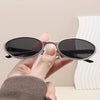 A hand holds a pair of black retro oval sunglasses with a silver metal frame against a white background.