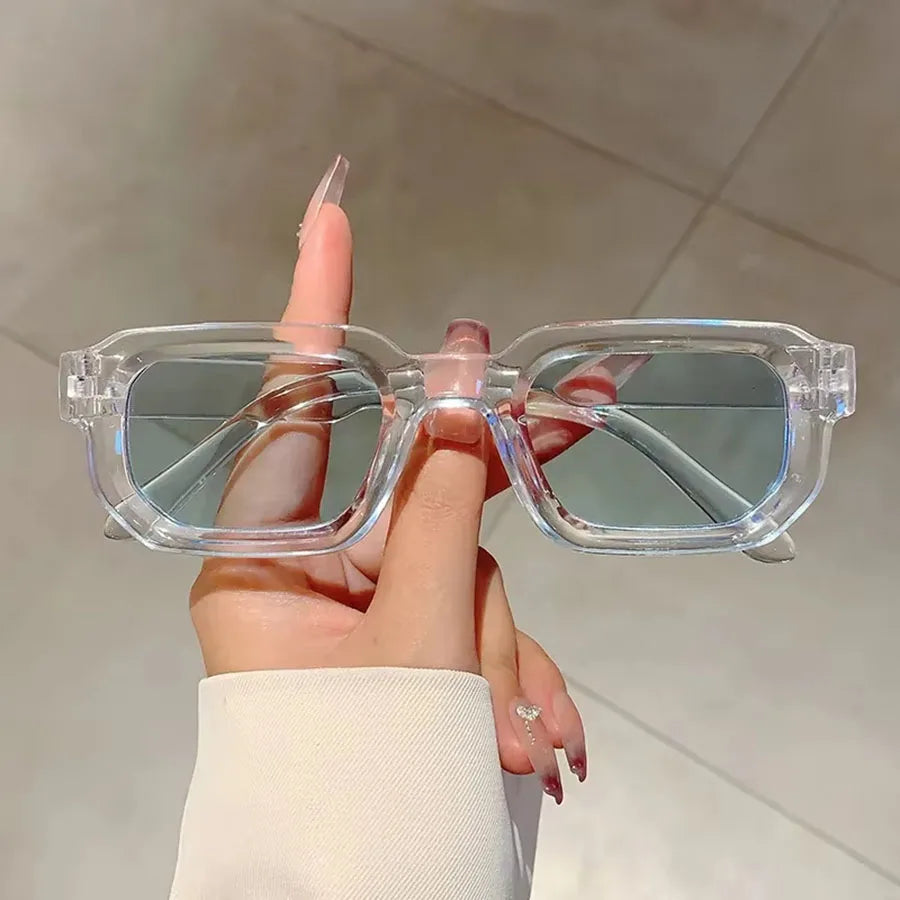 Une main tient des lunettes de soleil rectangulaires claires et rétro sur un fond clair.