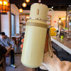 Une main tient une bouteille isotherme en acier inoxydable vert clair devant un arrière-plan flou d'un café intérieur.